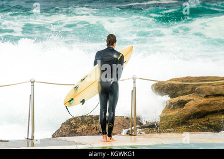 Un surfista maschio attende il momento giusto per entrare in acqua da Bronte Beach rock pool di Sydney, NSW, Australia in grandi condizioni surf Foto Stock