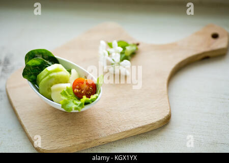 Piccolo bianco ovale ciotola riempita con materie cetrioli, peperoni, pomodori, gli spinaci e la lattuga, posto sul tagliere di legno Foto Stock