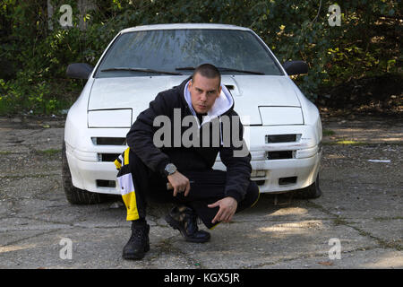 Un giovane uomo, gopnik inginocchiato accanto a un auto abbandonate Foto Stock