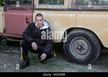 Un giovane uomo, gopnik inginocchiato accanto a un auto abbandonate Foto Stock