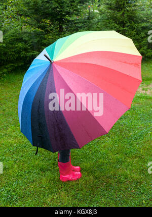 Bambina rosso da indossare stivali di gomma di nascondersi dietro ombrello coloratissimo sotto la pioggia Foto Stock