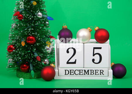 Close up di blocchetti di legno che mostra la data di calendario dicembre 25th, natale, seduti su uno sfondo verde con piccole decorate albero di natale accanto ad essa, sever Foto Stock