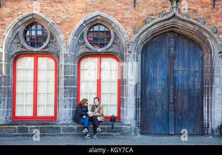 Bruges, Belgio - 11 Aprile 2017 - Due femmina felice Europa i turisti a sedersi su una vecchia chiesa della finestra sgabello godendo il loro snack su una strada di Bruges, essere Foto Stock