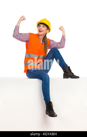 Felice giovane donna con elmetto giallo, gilet arancione riflettente, camicia Lumberjack, jeans, stivali neri, seduti sulla parte superiore con le braccia allungate e guardando u Foto Stock