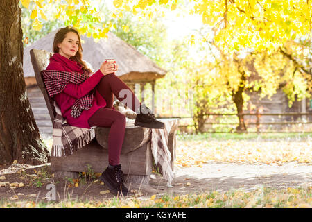 Sognando la donna sedersi sulla panchina in autunno park, coppa di ritegno e guardando lontano. outdoor shot Foto Stock