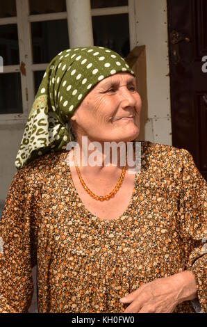Vecchia Signora orgoglioso, babushka, con sciarpa sul suo capo, collana e condire con pattern, è sorridente / ridere. in un piccolo villaggio in Tagikistan. Foto Stock