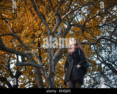 Una ragazza in un mantello nero passeggiate attraverso il giardino in autunno. gialla foglie degli alberi riempite la terra attorno a. Esso è caldo clima soleggiato. Foto Stock