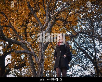 Una ragazza in un mantello nero passeggiate attraverso il giardino in autunno. gialla foglie degli alberi riempite la terra attorno a. Esso è caldo clima soleggiato. Foto Stock