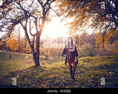 Una ragazza in un mantello nero passeggiate attraverso il giardino in autunno. gialla foglie degli alberi riempite la terra attorno a. Esso è caldo clima soleggiato. Foto Stock