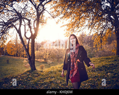 Una ragazza in un mantello nero passeggiate attraverso il giardino in autunno. gialla foglie degli alberi riempite la terra attorno a. Esso è caldo clima soleggiato. Foto Stock