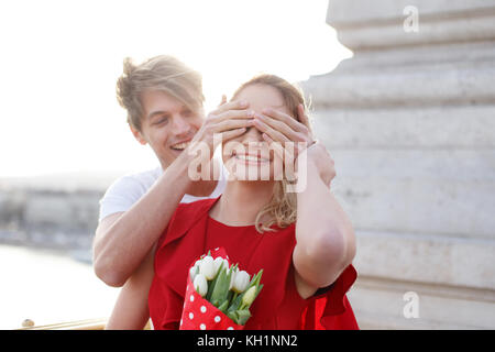 Giovane uomo nascondere girlfiends occhi, sorpresa sulla datazione, donna in abito rosso con bouquet Foto Stock