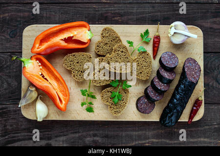 Morcillo (spagnolo black pudding, salsicce di sangue), taglio a fette, nero il pane di segale in una forma di cuore, pepe, aglio su una tavola di legno. La vista dall'alto su un Foto Stock