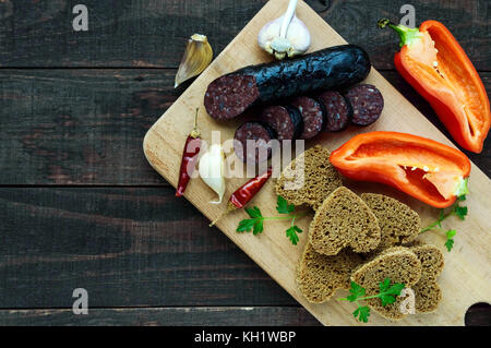 Morcillo (spagnolo black pudding, salsicce di sangue), taglio a fette, nero il pane di segale in una forma di cuore, pepe, aglio su una tavola di legno. La vista dall'alto su un Foto Stock