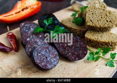 Morcillo (spagnolo black pudding, salsicce di sangue), taglio a fette, nero il pane di segale in una forma di cuore, pepe, aglio su una tavola di legno. close up. Un festi Foto Stock