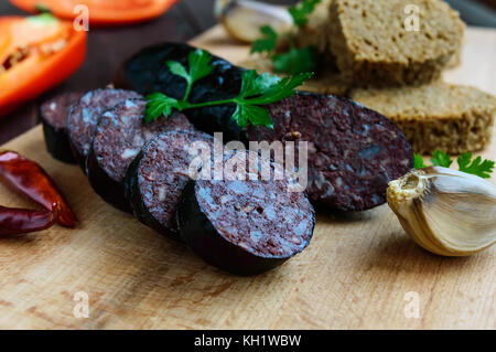 Morcillo (spagnolo black pudding, salsicce di sangue), taglio a fette, nero il pane di segale in una forma di cuore, pepe, aglio su una tavola di legno. close up. Un festi Foto Stock