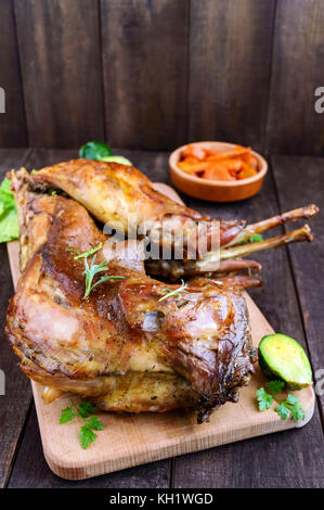 Arrosto di coniglio intero su una tavola di legno con carote al forno e cavolini di Bruxelles su uno sfondo scuro. Un pasto di festa. Foto Stock