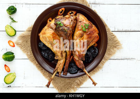 Arrosto di coscia di coniglio con prugne su una piastra di ceramica su sfondo chiaro. La vista superiore. menu per le vacanze di pasqua e natale. Foto Stock