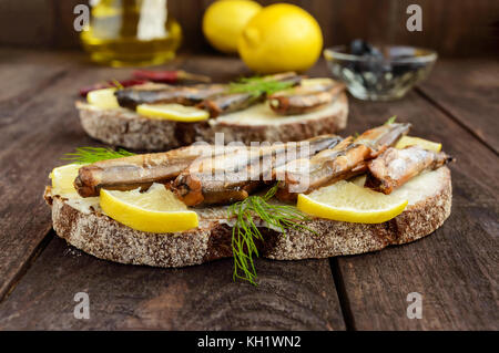 Sandwich con pesce affumicato capelin (spratti), limone sul nero pane di segale. snack al buio su un tavolo di legno. Foto Stock