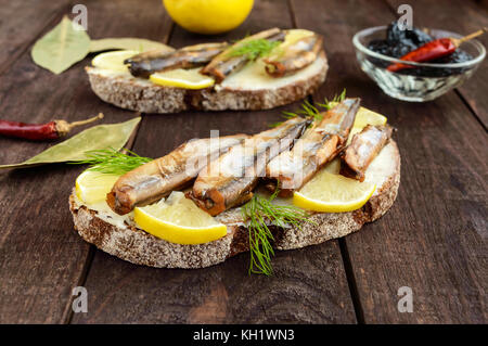 Sandwich con pesce affumicato capelin (spratti), limone sul nero pane di segale. snack al buio su un tavolo di legno. Foto Stock