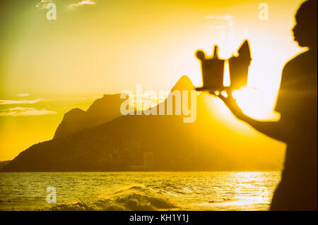 Brasiliano sfocati spiaggia trasporto fornitore caipirinha cocktail al arpoador con una vista al tramonto dei due fratelli di montagna in rio de janeiro, Brasile Foto Stock