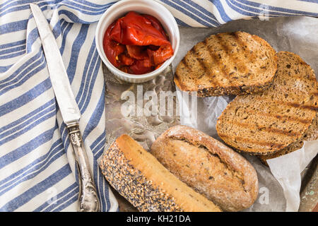 Pane integrale panini e pane grigliato fette, con arrosti di peperoni rossi nella ciotola e coltello. Foto Stock