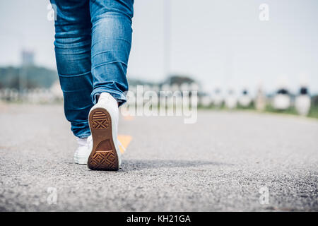 Close up donna indossare jean e bianco sneaker a camminare in avanti sulla highway road nella giornata di sole,da soli di viaggio o di persona che viaggia da sola nozione. Foto Stock