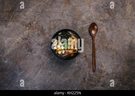 Giapponese zuppa di miso con tofu e il salmone in una ciotola nero su un vintage sfondo colorato con un cucchiaio di legno. vista verticale dall'alto. Foto Stock