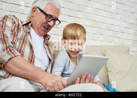Ritratto di felice senior uomo con tavoletta digitale per guardare video con il nipote entrambi ridendo seduti sul divano di casa Foto Stock
