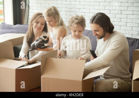 La famiglia felice di quattro con due bambini disimballaggio scatole in movimento nella nuova casa, sorridente i bambini aiutano i genitori con effetti personali in cartoni aperti seduta o Foto Stock