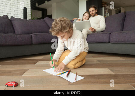 Ragazzo di età prescolare figlio di Disegno con matite colorate in album mentre i genitori usando il computer portatile sul lettino a sfondo, creative ragazzino giocando sul soggiorno Foto Stock