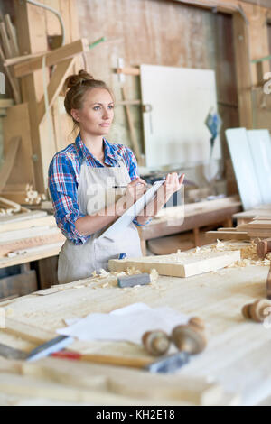 Ritratto di giovane donna negli appunti di contenimento e di rendere note durante la pianificazione di un nuovo progetto in una moderna officina per la lavorazione del legno Foto Stock