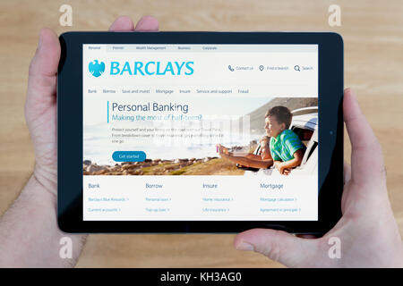 Un uomo guarda presso la Barclays Bank sito sul suo iPad dispositivo tablet, sparato contro un tavolo di legno top sfondo (solo uso editoriale) Foto Stock