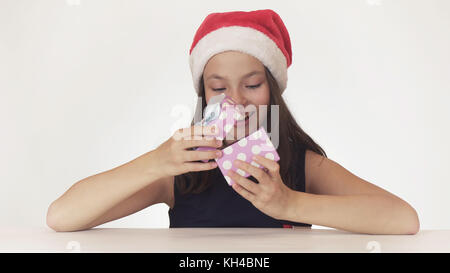 Bella ragazza teen in santa claus hat seduto e gioiosamente apre la confezione regalo su sfondo bianco Foto Stock