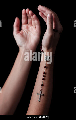 Mani femminili con le braccia aperte di pregare e di azienda rosario con croce o crocifisso. sfondo nero. donna con cristiana cattolica fede a mano Foto Stock