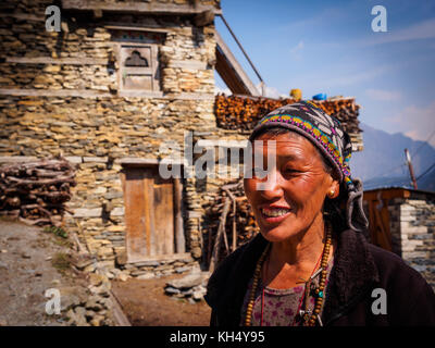 Weathered, il volto sorridente di una donna Manangi davanti al suo tradizionale casa di pietra in alto Pisang village, Circuito di Annapurna Trek, Nepal Foto Stock