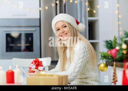 Una ragazza bionda in un berretto di Babbo Natale con un regalo in mano per c Foto Stock