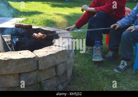 Due ragazzi sono la cottura alla griglia o alla brace salsicce polacco su un fuoco aperto buca nel loro cortile Foto Stock