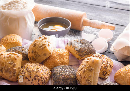 Tema di cottura immagine con panini fatti in casa decorata con semi di papavero, di sesamo, di lino e semi di girasole, e gli ingredienti per la loro realizzazione in background. Foto Stock