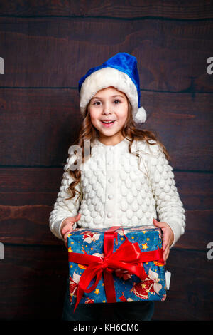 Piccolo felice sorridente ragazza con un regalo di Natale box.Concetto di Natale. Sorridente funny girl in Santa hat su sfondo di legno. Foto Stock