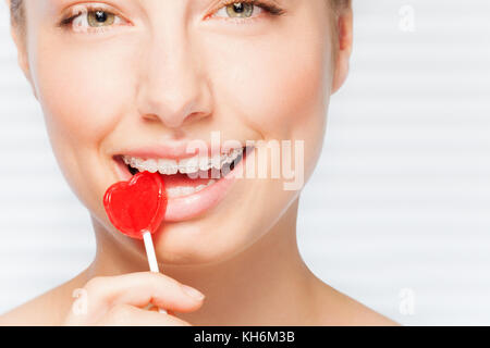 Ritratto di giovane e bella donna con bretelle dentali mordere off lecca-lecca rosso a forma di cuore Foto Stock