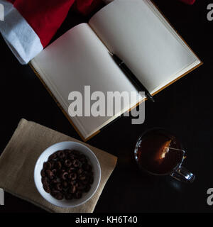 Pianificazione dei futuri sul Natale: un notebook con pagine vuote, una penna nera, santa hat, tazza di vetro con bustine di the, bianco ciotola di cioccolato anelli di cereali e mil Foto Stock