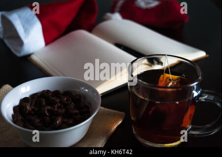 Pianificazione dei futuri sul Natale: un notebook con pagine vuote, una penna nera, santa hat, tazza di vetro con bustine di the, bianco ciotola di cioccolato anelli di cereali e mil Foto Stock