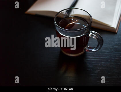 Un notebook con pagine vuote, una penna nera, tazza di vetro con bustine di the sulla scrivania scuro Foto Stock