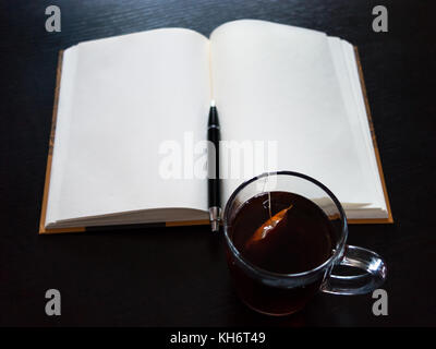 Un notebook con pagine vuote, una penna nera, tazza di vetro con bustine di the sulla scrivania scuro Foto Stock