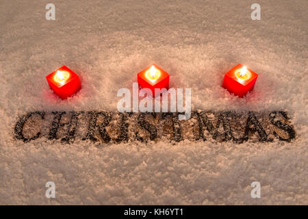 La parola natale in lettere di legno su un sfondo di neve illuminata da tre candele rosse. Foto Stock