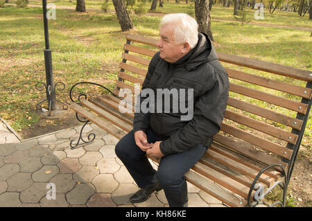 Il lone grey-haired uomo vecchio, in appoggio su una panca di legno in un parco su una soleggiata giornata autunnale. Foto Stock
