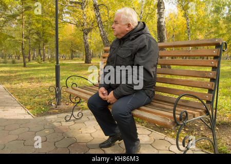 Il lone grey-haired uomo vecchio, in appoggio su una panca di legno in un parco su una soleggiata giornata autunnale. Foto Stock