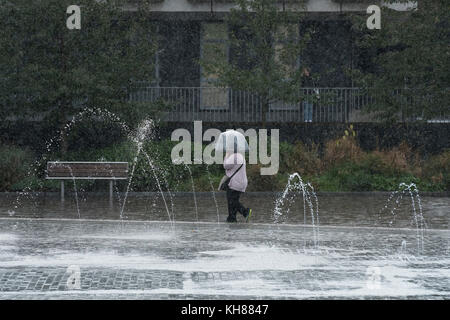 Donna che cammina in heavy rain, rifugiandosi sotto ombrellone, passando la piscina a specchio e fontane - Bradford City Park, West Yorkshire, Inghilterra, Regno Unito. Foto Stock