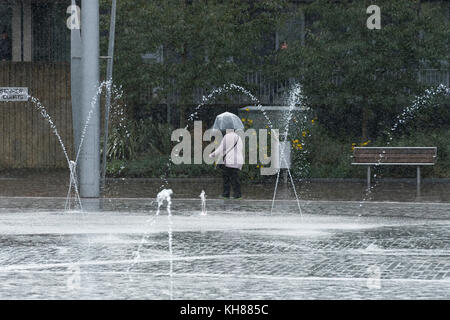Donna che cammina in heavy rain, rifugiandosi sotto ombrellone, passando la piscina a specchio e fontane - Bradford City Park, West Yorkshire, Inghilterra, Regno Unito. Foto Stock