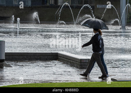 Giovane a piedi sotto la pioggia, rifugiandosi sotto ombrellone, passando la piscina a specchio e fontane - centro di Bradford City Park, West Yorkshire, Inghilterra, Regno Unito. Foto Stock